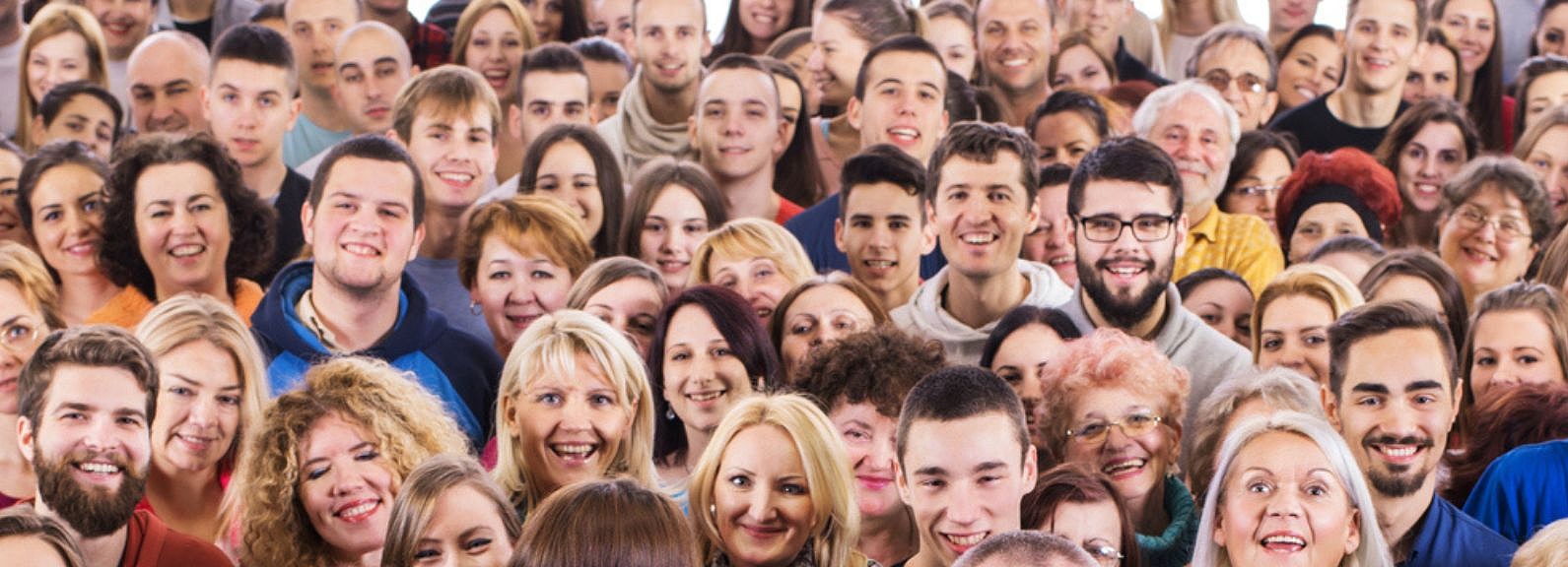Eine große, dichte Menschenmenge von Personen unterschiedlichen Alters und Geschlechts steht dicht beieinander und blickt in die Kamera. Viele Menschen lächeln und scheinen sich bei einer Veranstaltung oder einem Treffen zu befinden, dessen Hintergrund unscharf ist und auf einen Innenraum hindeutet. Die Aufnahme ist nah an der Menge, sodass die Gesichter im Vordergrund klarer erkennbar sind, während die hinteren Reihen verschwimmen. Die allgemeine Atmosphäre wirkt positiv und gemeinschaftlich.