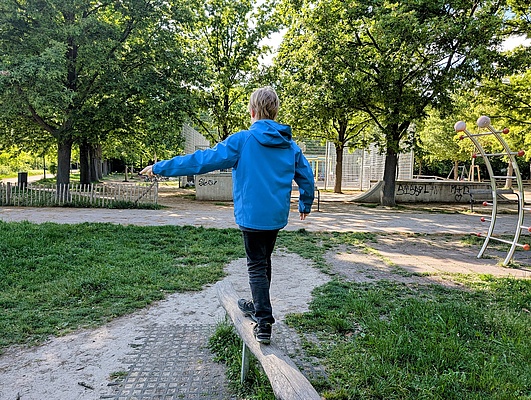 ein blonder Junge in blauer Jacke balanciert auf einem Balken auf einem Spielplatz