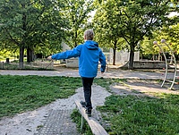 ein blonder Junge in blauer Jacke balanciert auf einem Balken auf einem Spielplatz