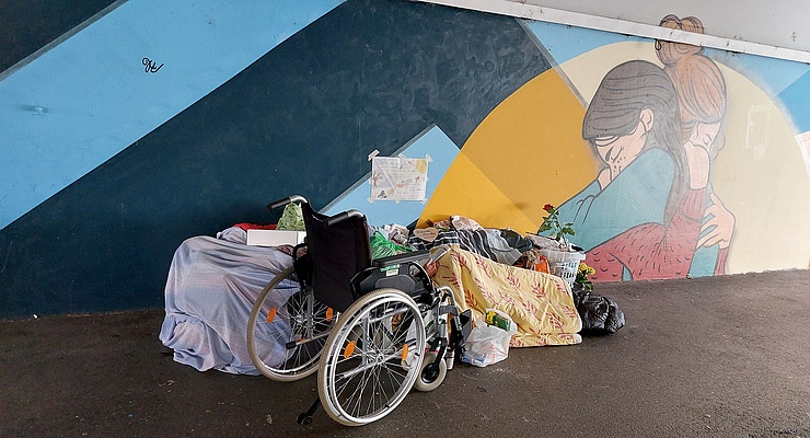 ein Rollstuhl, eine Decke und einige Plastiksäcke mit persönlichen Sachen in einer Unterführung. An der Wand dahinter ein Graffiti, das zwei Frauen zeigt, die sich umarmen