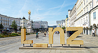Blick auf den Linzer Hauptplatz. Im Vordergrund eine Skulptur aus den Buchstaben LINZ, das I ist umgefallen, sodass eine Lücke entsteht