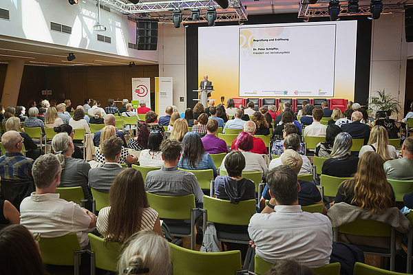 Publikum in Rückenansicht, Peter Schlaffer am Stehpult, er trägt kurze Haare und ein graues Sakko. Hinter ihm ist ein großer Screen zu sehen