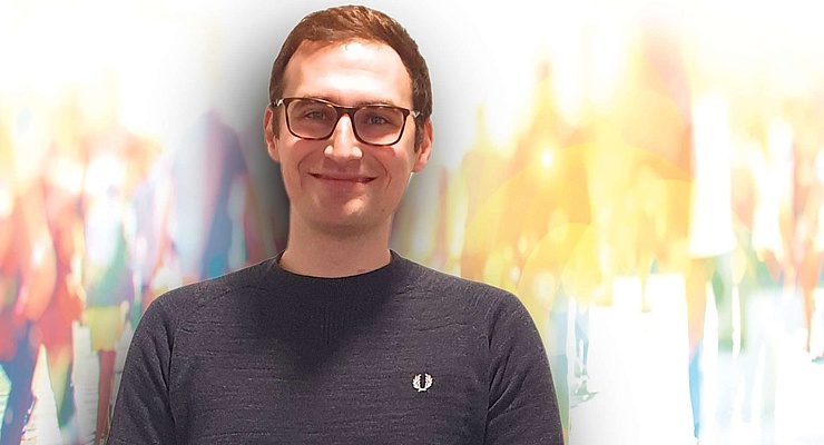 Ein lächelnder junger Mann mit Brille und einem dunkelblauen Pullover steht vor einem verschwommenen Hintergrund mit hellen, bunten Lichtern und schemenhaften Figuren, die Bewegung andeuten.