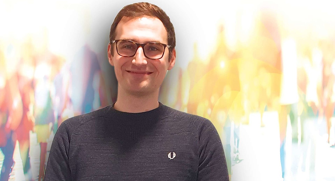 Ein lächelnder junger Mann mit Brille und einem dunkelblauen Pullover steht vor einem verschwommenen Hintergrund mit hellen, bunten Lichtern und schemenhaften Figuren, die Bewegung andeuten.