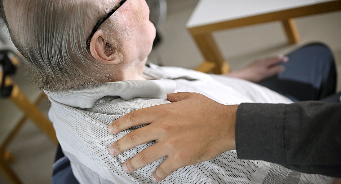 alter Mann im Pflegeheim, sitzend in Rückenansicht, graue Haare und Brille, die Hand eines jungen Mannes liegt auf seiner Schulter