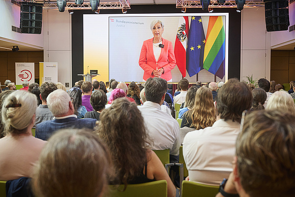 Publikum in Rückenansicht, auf einem großen Monitor ist Justizministerin Anna Sporrer zu sehen, sie trägt kurze graue Haare und einen lachsfarbenen Blazer. Neben ihr rechts sind die österreichische Flagge, die EU-Flagge sowie die Regenbogenflagge.   
