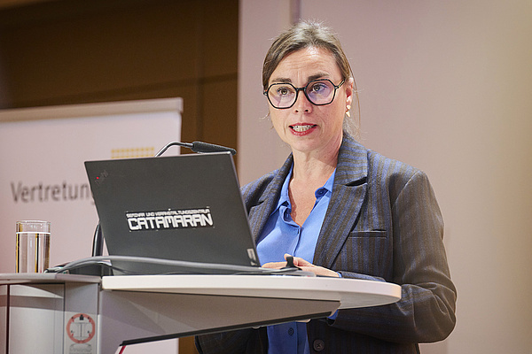 am Stehpult Karin Rowhani-Wimmer, sie trägt ihre dunklen Haare zusammengebunden und eine Brille sowie einen grauen Blazer