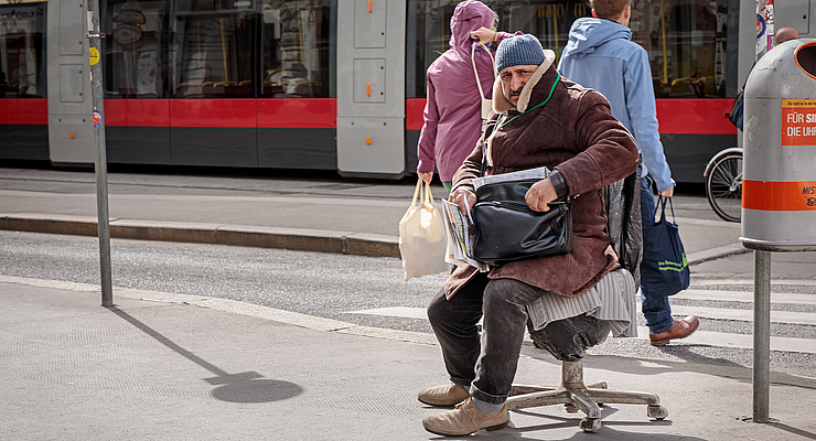 ein älterer Straßenzeitungsverkäufer sitzt an einer Straßenbahnhaltestelle auf einem Drehstuhl. Am Schoß hält er eine schwarze Tasche. Im Hintergrund überqueren zwei Menschen die Straße, eine Straßenbahn steht am gegenüberliegenden Gleis