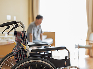 Im Pflegeheim, eine Person sitzt im Bett, der Rollstuhl steht zu weit entfernt zum Aufstehen