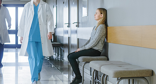 eine blonde junge Frau in grauem Pullover und schwarzer Hose sitzt auf einem Warteplatz im Krankenhaus. Ihr Blick ist resigniert. Eine Ärztin in weißem Kittel und türkiser Hose geht vorüber