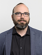 Thomas Berghammer in graublauem Sakko und schwarzem Hemd, er trägt kurze Haare und eine Brille