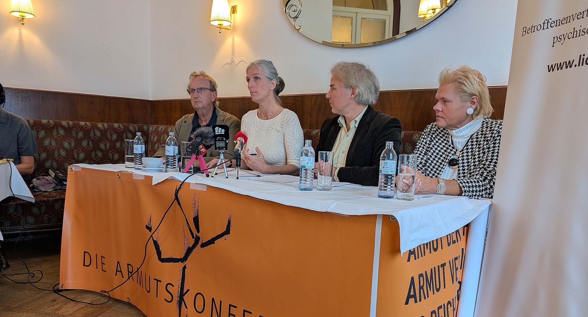 Podium mit zwei Männern und zwei Frauen, davor ein oranges Plakat mit dem Logo der Armutskonferenz, ein auf einem Bein balancierendes Strichmännchen. Gerade spricht Gerlinde Heim, eine Frau mit langen grauen Haaren, sie trägt eine beige Bluse 