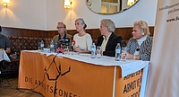 Podium mit zwei Männern und zwei Frauen, davor ein oranges Plakat mit dem Logo der Armutskonferenz, ein auf einem Bein balancierendes Strichmännchen. Gerade spricht Gerlinde Heim, eine Frau mit langen grauen Haaren, sie trägt eine beige Bluse 