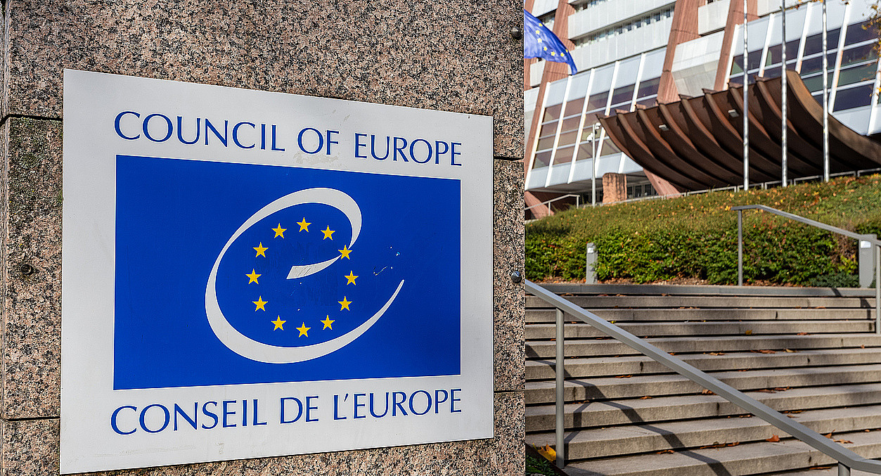 Stufen zum Gebäude des Europarats, links davon das blaue Logo des Europarats auf einem Schild an einer Mauer